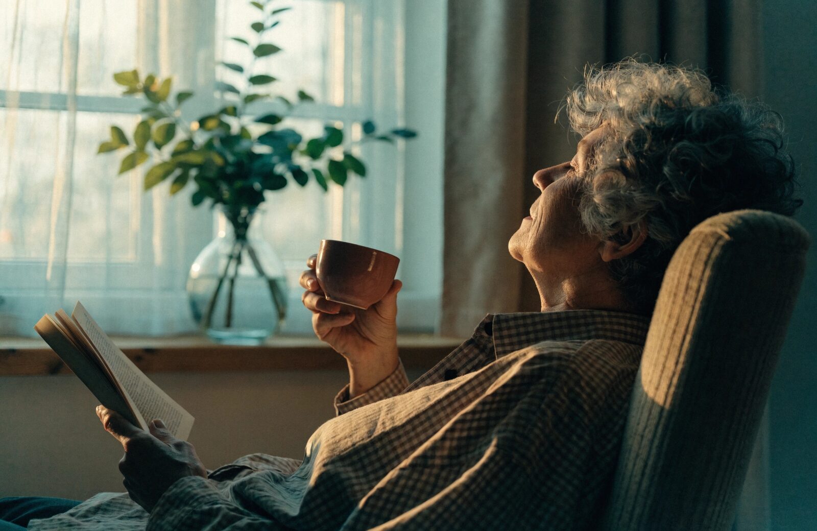 An older adult relaxing in an armchair with a book by the window.